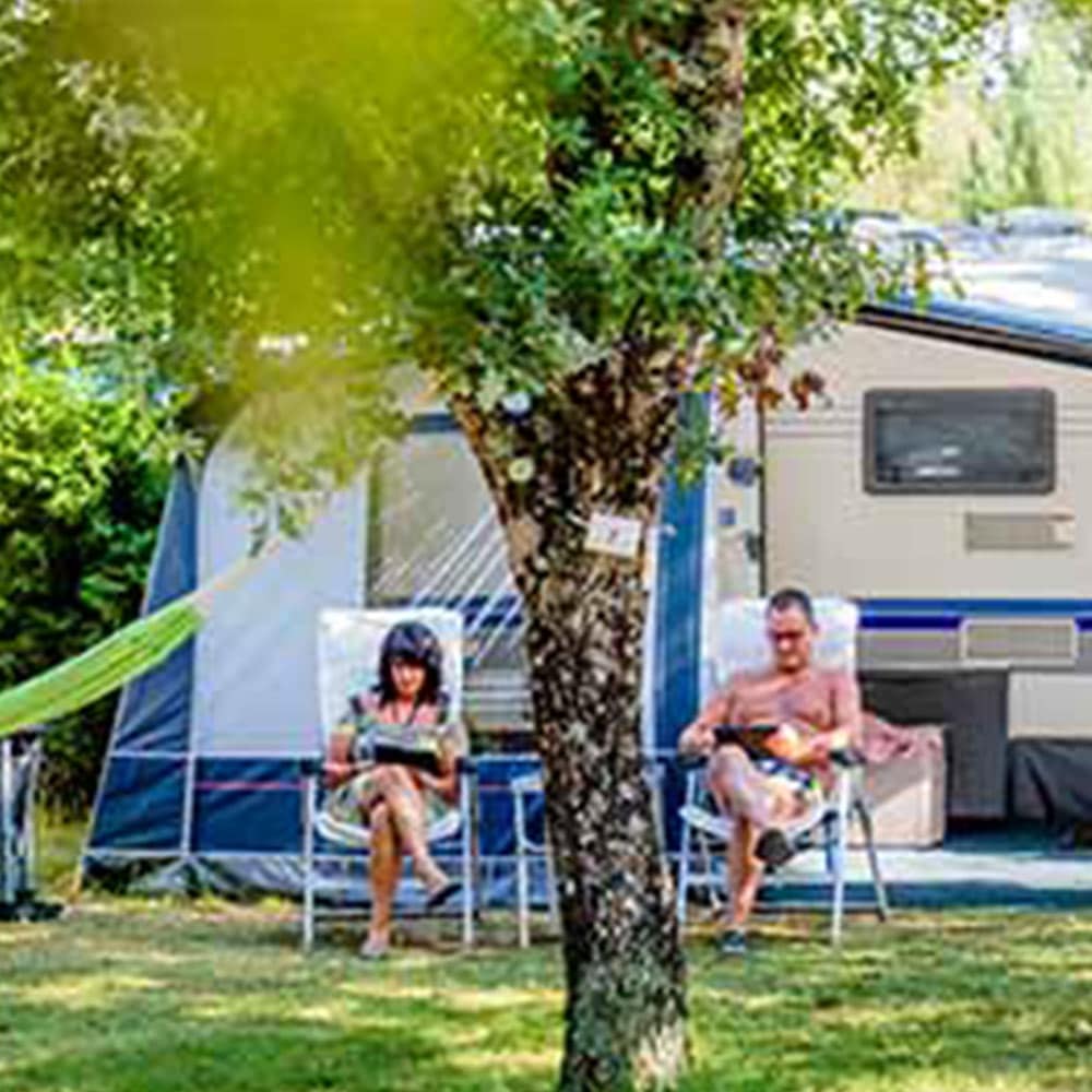 Couple se relaxant sur des chaises de camping devant une caravane et une tente. La femme lit, l'homme regarde une tablette près d'un arbre.