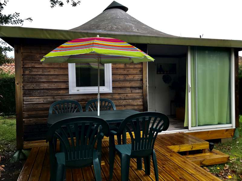 Petit chalet en bois avec terrasse, mobilier de jardin vert et parasol rayé sous un toit en forme de yourte.