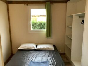 Chambre avec lit double, draps gris, oreillers blancs, fenêtre avec rideau vert et étagères de rangement ouvertes.