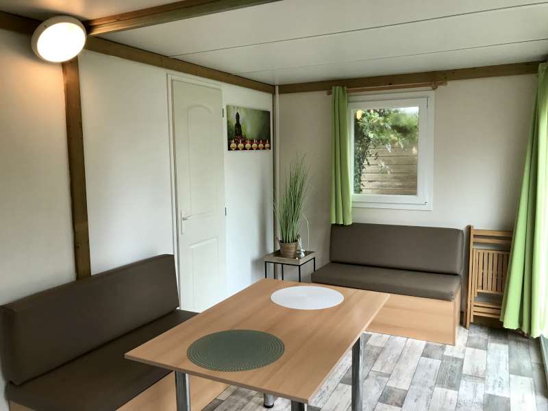 Intérieur lumineux d'un hébergement avec table, banquettes marron, rideaux verts et décoration zen épurée.